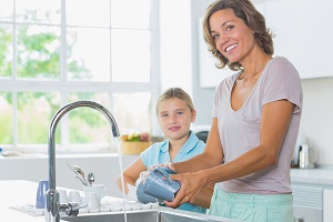 Preteen girl standing washing dishes at the kitchen sink with her mother in front of running faucet
